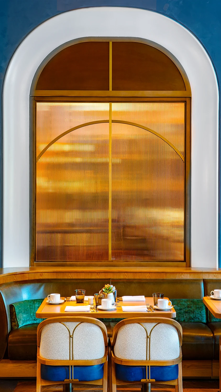 An arched gold-stained glass window behind a wooden dinner table set for 4 persons seated at a set of chairs and an upholstered booth